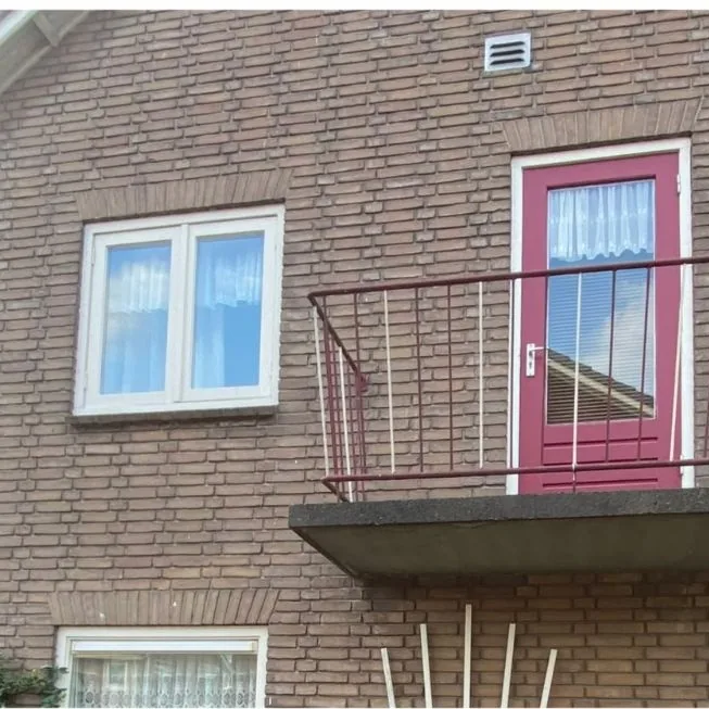 Balkon met een rood hekwerk en een rode deur in een bakstenen gevel, vóór het werk